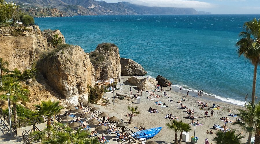 Playa de Burriana in Nerja: golden sand beach framed by dramatic ochre cliffs and palm trees, with sunbathers under thatched parasols and the deep blue Mediterranean stretching toward the Sierra de Almijara mountains