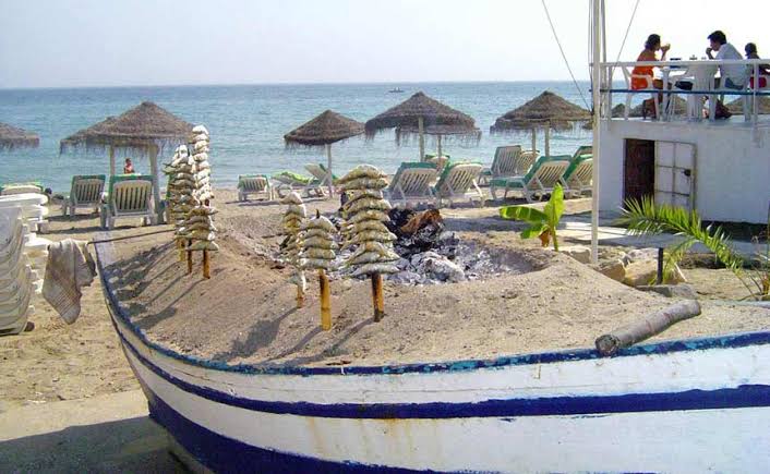 Espetos de sardinas grilling on a traditional jábega fishing boat at Pedregalejo beach, with thatched parasols and sunbathers behind