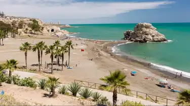 Peñón del Cuervo beach with its iconic offshore rock, palm trees and dark sand on a sunny day