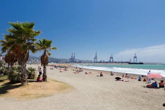 Playa de San Andrés in Málaga: tall palm trees on the left edge a wide sandy beach dotted with sunbathers and a red-and-white striped parasol, with the cranes of the port of Málaga visible across the bay